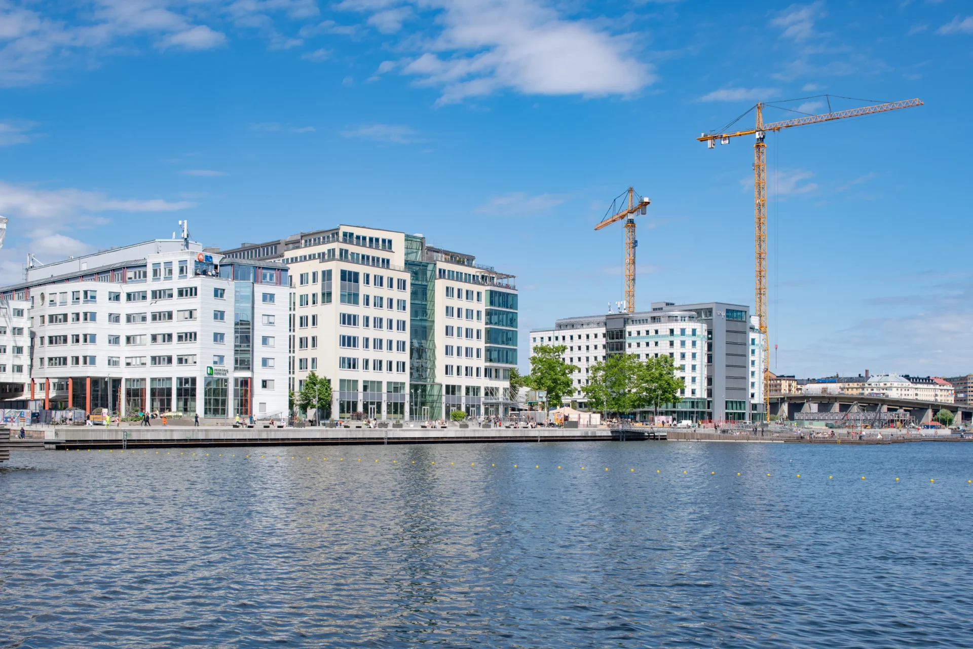 Vy över en stad från vattnet, med höga byggnader och lyftkranar i bakgrunden i starkt solljus.