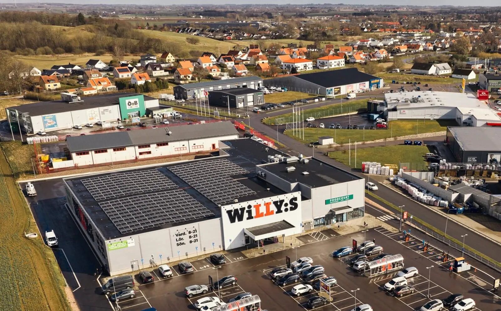 Närmare flygvy över en handelsplats fylld av parkerade bilar och omgivande grönområden.
